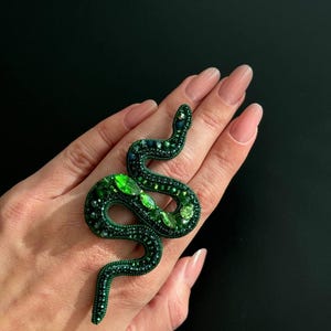 May include: A green snake-shaped brooch with emerald green and dark green rhinestones. The brooch is on a hand.