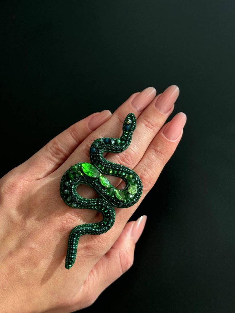 May include: A green snake-shaped brooch with emerald green and dark green rhinestones. The brooch is on a hand.