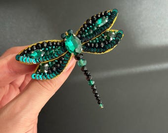 May include: A beaded dragonfly brooch with emerald green and black sequins. The wings are outlined in gold thread, and the body features a large green gemstone. The dragonfly's tail is made of black and green beads. A unique accessory.