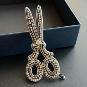 Puede incluir: Broche de tijeras plateadas, intrincadamente detallado con filas de pequeños cristales brillantes. Las hojas y los mangos de las tijeras están adornados con los cristales, y los lazos de las tijeras tienen detalles en negro. Un solo cristal negro cuelga de la parte inferior.