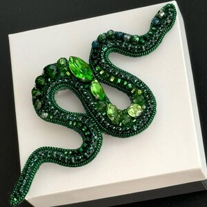 May include: A green snake brooch made of beads and rhinestones. The snake is curled up on a white surface.