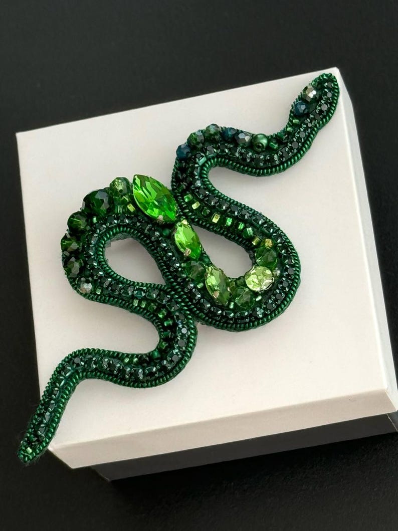 May include: A green snake brooch made of beads and rhinestones. The snake is curled up on a white surface.