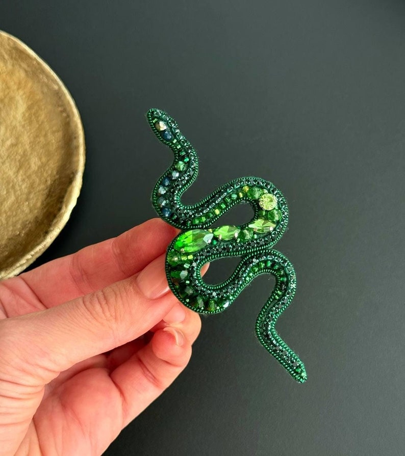 May include: A green beaded snake brooch with a coiled body and a long tail. The snake is made of green beads and rhinestones.