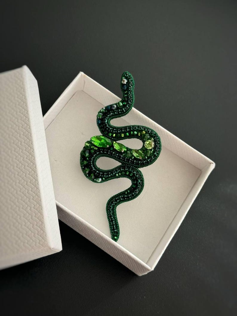 May include: A green beaded snake brooch with green and blue accents. The brooch is in a white box.