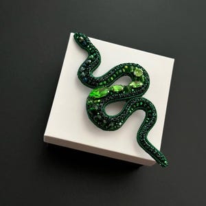 May include: A green beaded snake brooch with emerald green accents. The brooch is on a white square box.