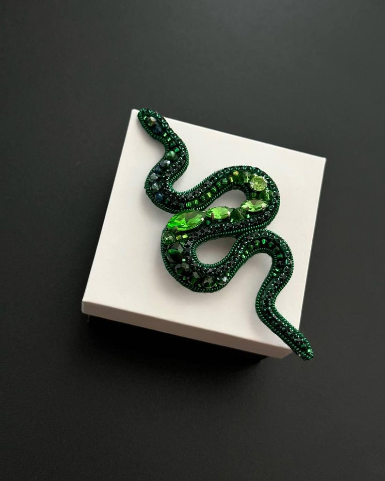 May include: A green beaded snake brooch with emerald green accents. The brooch is on a white square box.