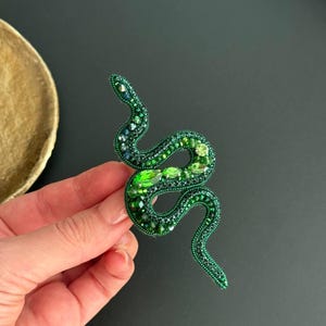 May include: A green beaded snake brooch with emerald green accents. The snake is curled in a S shape.