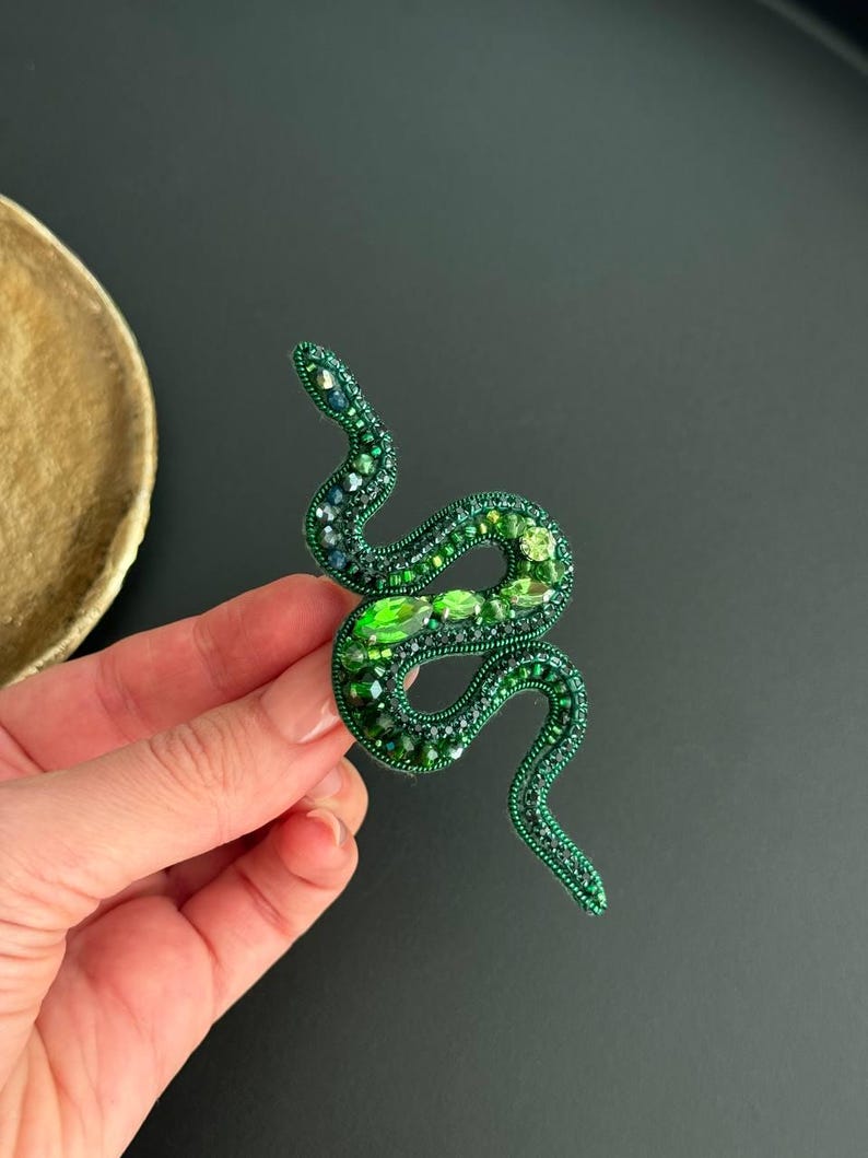 May include: A green beaded snake brooch with emerald green accents. The snake is curled in a S shape.