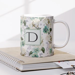 May include: White ceramic mug with a floral pattern and the letter "D" in a square frame. The mug is sitting on a spiral bound notebook.