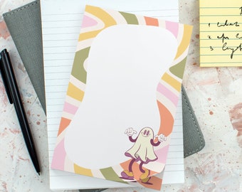 May include: A spiral-bound notepad featuring a central white area surrounded by a wavy, multi-colored border of pink, yellow, and olive green, with a cartoon ghost character at the bottom, placed atop a gray notebook and a yellow sticky note.