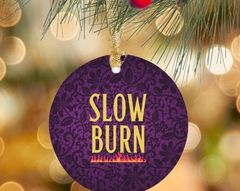 May include: Purple round Christmas ornament with a black lace pattern and gold text that reads "SLOW BURN" with a small orange flame graphic below the text.