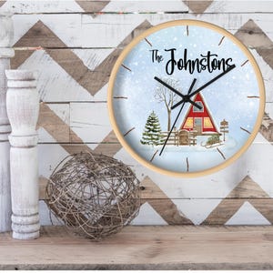 May include: A round wooden clock with a white background and a winter scene. The clock face has black hands and black Roman numerals. The scene features a red cabin with snow on the roof and a snowy landscape. The text "The Johnstons" is printed on the clock face.