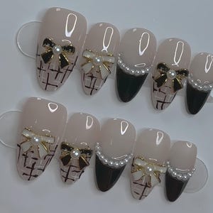 May include: Set of ten false nails with a pink base and black tips. Each nail is decorated with a white and gold bow, pearl accents, and a black and white checkered pattern.