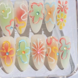 May include: A set of ten false nails with a white base and colorful flower designs. The flowers are painted in shades of pink, blue, green, and yellow.