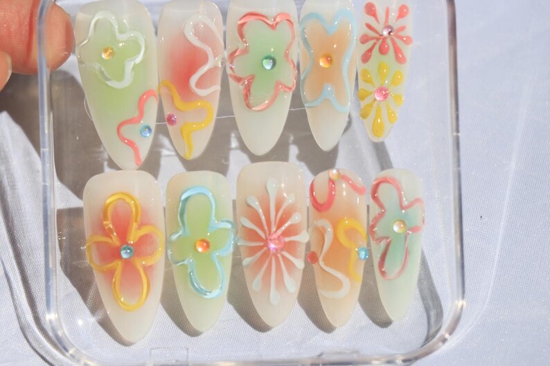 May include: A set of ten false nails with a white base and colorful flower designs. The flowers are painted in shades of pink, blue, green, and yellow.