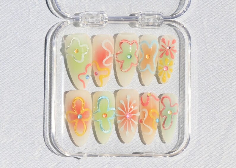 May include: A clear plastic case with ten false nails. Each nail is painted a light yellow and has a different floral design in pastel colors.