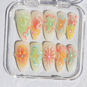May include: A set of ten false nails with a clear base and colorful floral designs. The designs include flowers, swirls, and dots in shades of pink, green, blue, and yellow.