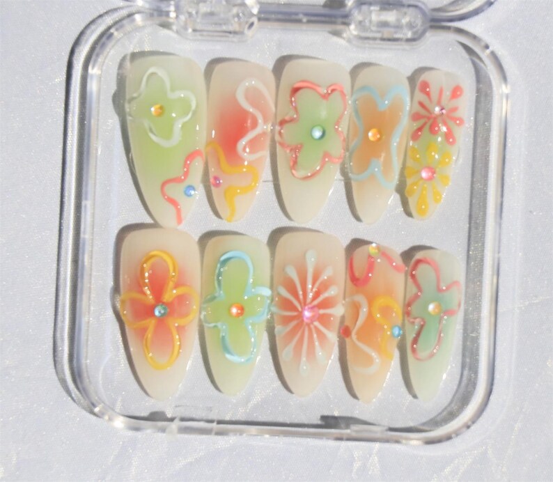 May include: A set of ten false nails with a clear base and colorful floral designs. The designs include flowers, swirls, and dots in shades of pink, green, blue, and yellow.