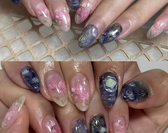 May include: Close-up of two sets of manicured nails. Each nail features a clear base with intricate floral designs in pink, white, and green. Some nails have a dark blue and white design. The nails are oval-shaped.