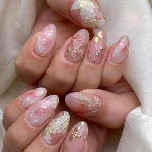 May include: Close-up of a hand with pink and white oval-shaped nails decorated with gold floral designs. The nails are painted with a light pink base and have delicate gold flowers and leaves on top.