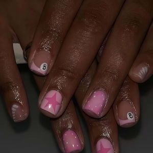 Puede incluir: Uñas manicuradas con un diseño rosa y blanco. Algunas uñas presentan una estrella rosa, otras lunares blancos y dos uñas muestran el número 8. Las uñas tienen un acabado brillante.