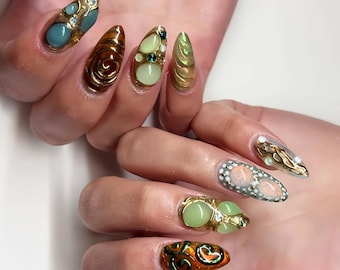 Puede incluir: Primer plano de uñas largas en forma de almendra con diseños intrincados. Las uñas presentan una variedad de colores, incluyendo dorado, turquesa, marrón y verde, con adornos como gemas y patrones arremolinados. La estética general es artística y detallada.
