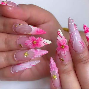 Puede incluir: Un conjunto de uñas artificiales largas y puntiagudas. Las uñas están pintadas en un color base rosa claro con varios diseños, incluyendo flores rosas, puntas blancas y detalles dorados. Algunas uñas presentan adornos florales en 3D y purpurina.