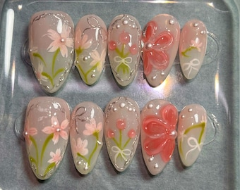 May include: Ten oval-shaped false nails with floral designs. The nails are painted in a light pink colour with white and green floral accents. Some of the nails have a 3D flower design.