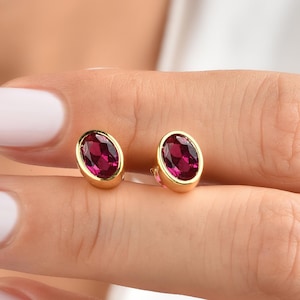 Könnte beinhalten: Ein Paar goldfarbene Ohrstecker, jeweils mit einem ovalen, tiefroten Edelstein. Die Ohrringe haben ein schlichtes, elegantes Design, das die leuchtende Farbe der Steine hervorhebt. Ein klassisches Schmuckstück.