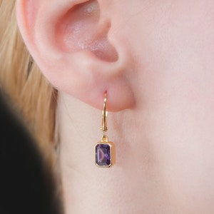 May include: A gold earring featuring a rectangular, deep purple gemstone. The earring is a leverback style, with the gemstone set in a gold bezel. The earring is shown on a person's ear.