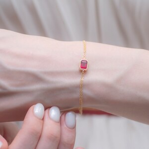 May include: A delicate gold chain bracelet with a single red gemstone in a square setting.