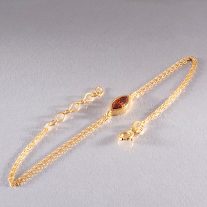 May include: A gold-coloured bracelet with a central marquise-cut, reddish-brown gemstone. The bracelet has a double chain design and an adjustable length with a lobster clasp closure. The gemstone is set in a gold bezel.