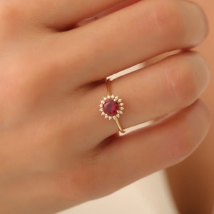 Könnte beinhalten: Ein goldener Ring mit einem roten Edelstein, der von kleineren weißen Edelsteinen umgeben ist.