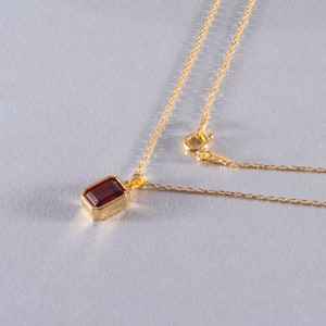 May include: A gold necklace with a rectangular, deep red gemstone pendant. The pendant is set in a gold bezel. The delicate gold chain is connected to a clasp. The jewelry is displayed on a light gray surface.