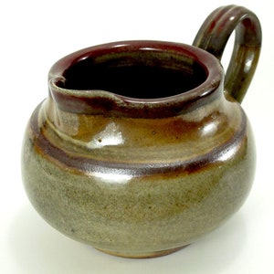 May include: A small, brown and green ceramic creamer with a rounded body and a single handle. The creamer has a brown rim and a brown stripe around the middle.