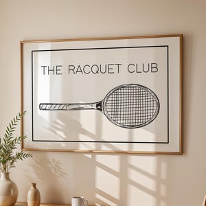 May include: A framed print with a black and white illustration of a tennis racket and the text "THE RACQUET CLUB". The print is hanging on a beige wall above a wooden table with a vase of green plants.
