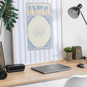 May include: A framed print with a blue and white striped background featuring a box of matches with a seashell design. The print is hanging on a wall above a desk with a laptop, a lamp, and a potted plant.