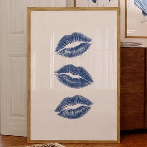 May include: A framed print featuring three blue lip prints on a white background. The artwork is displayed in a room with other framed art pieces, a wooden cabinet, and a coffee table.