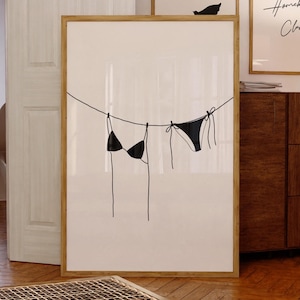 Può includere: Un disegno in bianco e nero di un top e un fondo bikini appesi a uno stendino. Il disegno è su sfondo bianco ed è incorniciato in una cornice di legno chiaro.