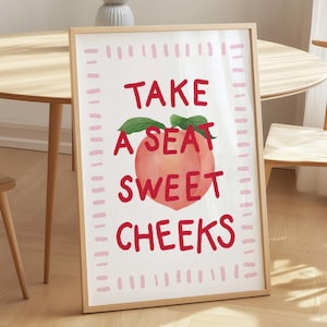 Könnte beinhalten: Gerahmter Kunstdruck mit den Worten "TAKE A SEAT SWEET CHEEKS" in Rot, mit einer Pfirsich-Illustration. Der Druck befindet sich in einem hellen Holzrahmen und lehnt an einer Wand. Ein runder Holztisch und ein Stuhl sind im Hintergrund.