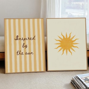 Könnte beinhalten: Zwei rechteckige gerahmte Wandbilder. Das linke Bild zeigt vertikale gelbe und cremefarbene Streifen mit dem Text "Inspired by the sun" in brauner Schreibschrift. Das rechte Bild zeigt eine gelbe Sonnenillustration auf cremefarbenem Grund.