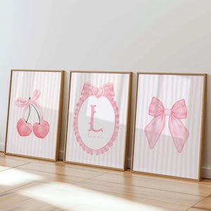 Op de afbeelding: Drie ingelijste kunstprints met roze aquarelontwerpen. De prints tonen een paar kersen met een strik, een roze strik en een roze ovaal met de letter "L" en de naam "Lisa". De achtergrond is roze en wit gestreept.