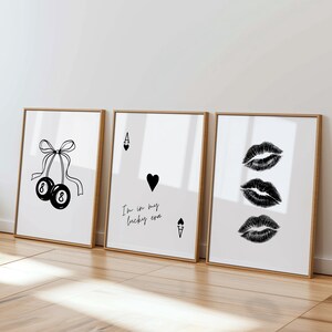 May include: Three framed art prints with a minimalist design. One print features two black 8-ball pool balls tied with a bow. Another shows an Ace of Hearts card with the text "I'm in my lucky era." The third print displays three sets of black lips.