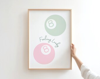 May include: A framed print featuring two stylized eight balls, one in pastel green and the other in pastel pink. The words "Feeling Lucky" are written in a script font between the two balls. The frame is a light wood color.
