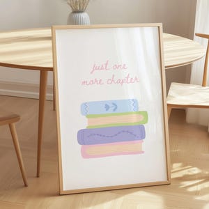May include: A framed vertical wall art print featuring the text "just one more chapter" in pink script above a stack of four illustrated books in light blue, green, purple, and pink colors with simple decorative line patterns on their covers.