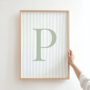 Puede incluir: Un cuadro enmarcado con la letra "P" en un tono verde suave. El fondo presenta rayas verticales en un tono similar. El marco es de madera clara.