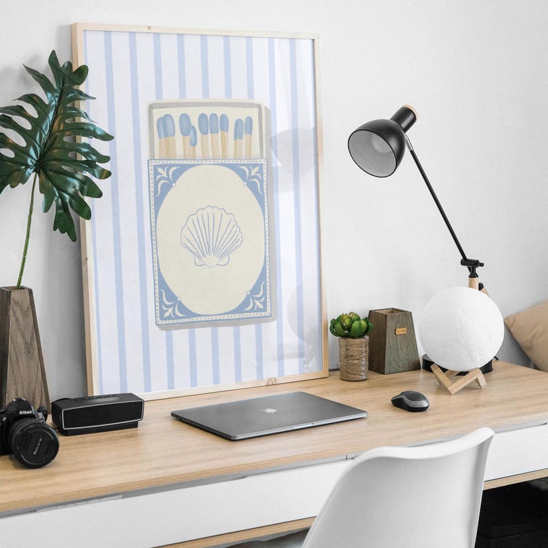 May include: A framed print with a blue and white striped background featuring a box of matches with a seashell design. The print is hanging on a wall above a desk with a laptop, a lamp, and a potted plant.