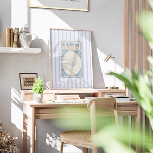 May include: A light wood desk with a chair and a framed print of a blue and white matchbox with a seashell design. The print is hanging on a light blue wall with white stripes. A small potted plant sits on the desk.
