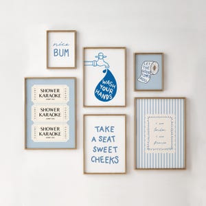 May include: A collection of framed art prints with gold frames and blue and white designs. The prints feature phrases like "nice BUM", "Wash Your Hands", and "Take a Seat Sweet Cheeks". Other prints include "Shower Karaoke" tickets and a striped design.