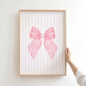 May include: A framed watercolor print featuring a large pink bow on a striped background of pink and white. The bow is centered and has a soft, delicate appearance. The frame is a light wood color.